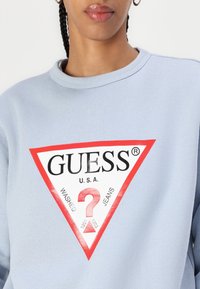 Felpa azzurra chiara con un grande logo triangolare rosso e bianco con "GUESS" e altro testo in nero. Texture morbida, design con collo rotondo.