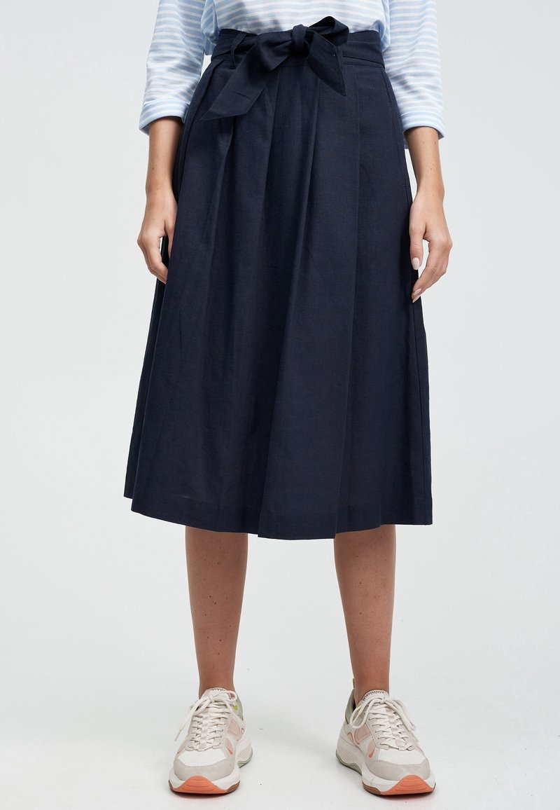 Mörkblå, knälång kjol gjord av texturerat tyg med en rosett vid midjan och veckad design. Matchas med ljusa sneakers.