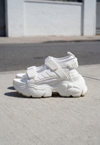 Sandales de sport blanches épaisses avec semelles texturées épaisses et brides Velcro réglables sur un trottoir en béton en plein jour.