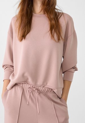 Femme portant un sweat ample rose poussiéreux et un pantalon de survêtement assorti avec cordon, mains dans les poches, vue du cou aux hanches.