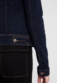 Veste en denim bleu foncé avec des coutures orange, dotée d'un poignet boutonné et d'un détail de bouton angulaire unique près de l'ourlet. Pantalon noir avec poche.