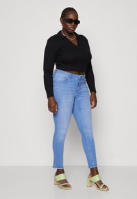 Black cropped long-sleeve top, light blue skinny jeans with frayed hems, and green heeled sandals with two wide straps. Simple silver chain accessory.