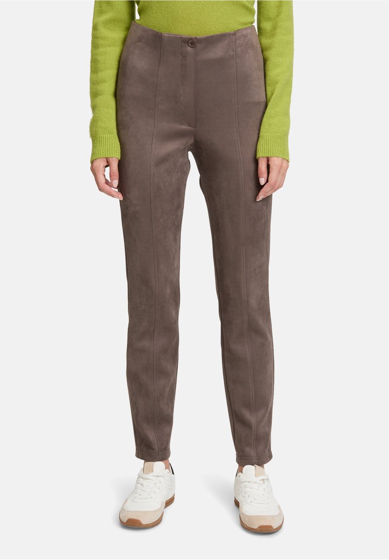 Vrouw in smal passende bruine suède broek, witte sneakers en een limoengroen langemouwentrui, staande tegen een effen achtergrond.