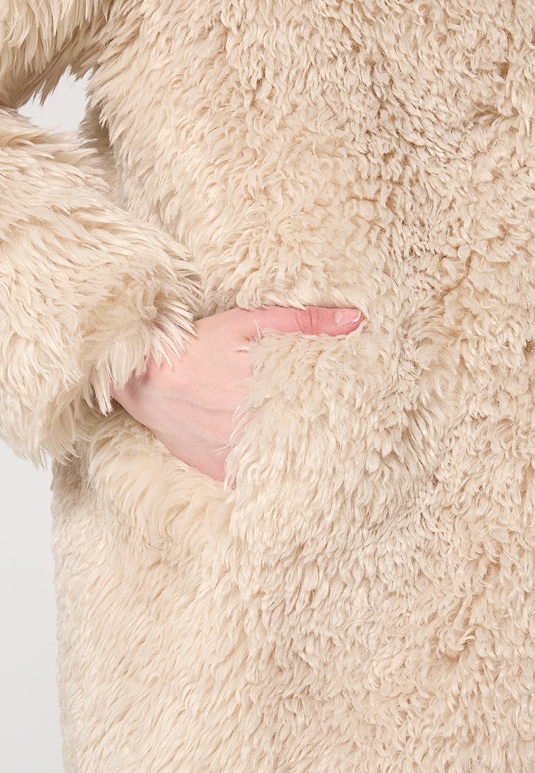 Beige Kunstpelz-Oberbekleidung mit einer weichen, strukturierten Oberfläche. Verfügt über eine Seitentasche; der Stoff hat ein volles, plüschiges Aussehen.