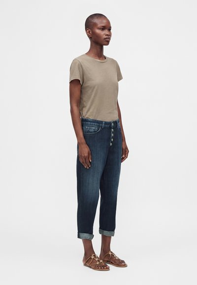 Personne debout portant une chemise beige à manches courtes, un jean foncé retroussé avec des boutons visibles, et des sandales marron sur un fond clair uni.