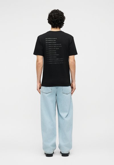 Άνδρας με σγουρά μαλλιά που φοράει μαύρο μπλουζάκι με λευκό κείμενο στην πλάτη και ανοιχτό μπλε φαρδιά τζιν, στέκεται μπροστά σε απλό λευκό φόντο.