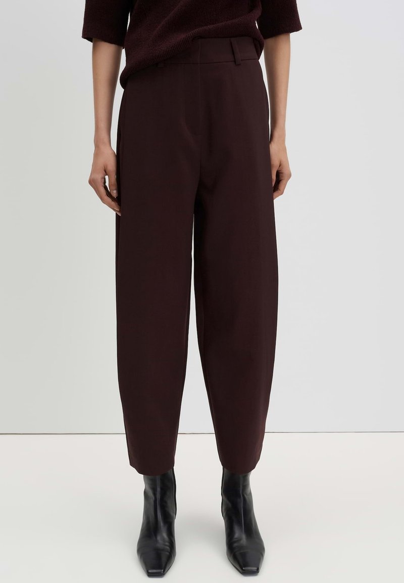 Person wearing high-waist dark brown tapered pants and black ankle boots standing against a plain light background.