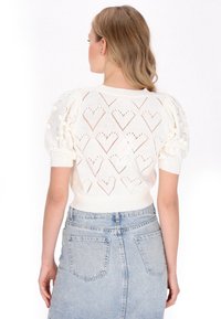 Hvid strikket sweater med puffærmer, der har hjerteformede udskæringer og strukturerede bobble-detaljer, parret med en let denimnederdel.