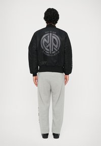 Jacheta bomber neagră are un logo mare și text pe spate, cu manșete și tiv ribbed, asortată cu pantaloni de trening gri deschis.