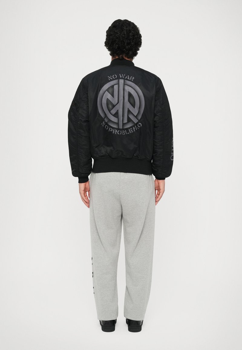 Jacheta bomber neagră are un logo mare și text pe spate, cu manșete și tiv ribbed, asortată cu pantaloni de trening gri deschis.