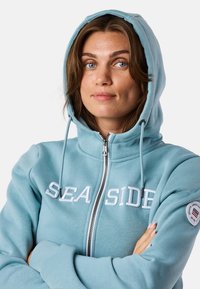 Lichtblauwe zip-up hoodie met een zachte textuur, met een wit geborduurd logo "SEASIDE" en een ronde logo patch op de mouw.