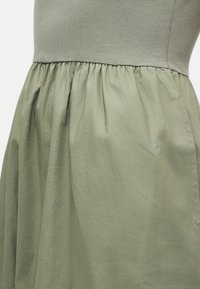 Κοντινό πλάνο ενός ελιά πράσινου φορέματος με εφαρμοστό πάνω μέρος και λεπτομέρεια φούστας με πιέτες και ρέον ύφασμα.