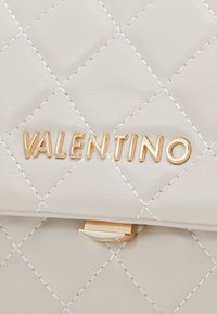 Bolso de mano de cuero beige acolchado con herrajes dorados; presenta un patrón de diamantes cosido y el logotipo "VALENTINO" en relieve en la parte frontal.