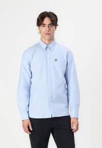 Chemise boutonnière bleu clair avec une poche poitrine, arborant un petit logo en forme de papillon jaune, fabriquée en tissu doux avec une texture lisse.