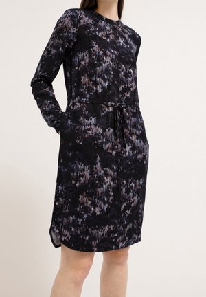 Femme portant une robe noire à manches longues, longueur genou, avec des motifs abstraits violets et gris et une taille à cordon, mains dans les poches.