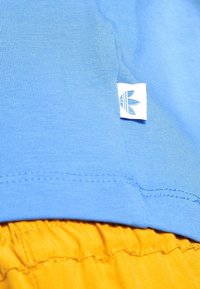 Blå bomullströja med en liten vit etikett som har en lövlogotyp, i kombination med gult tyg som syns lite under.