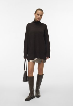 Vrouw in een oversized donkere coltrui, metalen minirok, kniehoge bruine laarzen en een zwarte handtas.