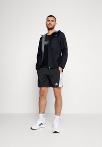Chaqueta ligera negra con acentos grises, camiseta negra, pantalones cortos negros con patrones laterales blancos y zapatillas deportivas blancas.