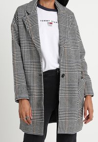 Svart- och vitrutigt kavaj med notched krage, oversize passform, enkel knappstängning och texturerat tyg. Lager på en vit T-shirt.