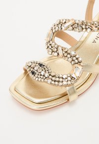 Gullhelete sandal med firkantet tå, med en surrende stropp dekorert med klare og svarte rhinestones og en metallisk finish.