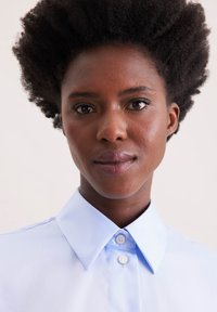 Camicia azzurra chiara con colletto e texture liscia, dotata di bottoni e un design strutturato, indossata da una persona con capelli scuri e con texture.