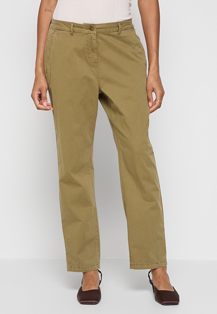 Frau in hoch taillierten, geraden khakifarbenen Hosen mit seitlichen Taschen und braunen, eckigen Schuhen, die vor einem hellgrauen Hintergrund steht.