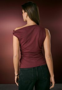 Bordeaux, off-shoulder bluse med fitted design, der har indsamlet detaljer på ryggen, parret med mørke jeans. Glat stoftekstur.