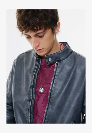 Sort læderjakke med stålkrave og sølvhardware, båret over en bordeaux strikket sweater. Et enkelt vedhæng hænger fra en brun snor.