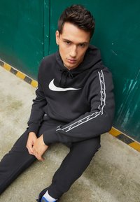 Felpa nera abbinata e pantaloni della tuta realizzati in misto cotone, con il logo Nike bianco sul petto e strisce laterali con piccoli loghi ripetuti.