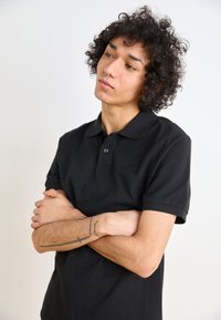Black polo shirt with a collar and short sleeves, featuring a subtle logo on the chest. The fabric appears smooth and slightly textured.