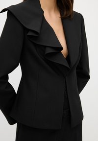 Blazer noir cintré avec grand revers à volants porté sur un top en dentelle décolleté, présenté de la clavicule jusqu'aux hanches sur un fond uni.