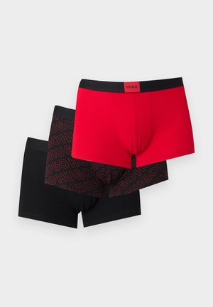 Homme athlétique debout pieds nus en boxer rouge avec une ceinture noire sur fond uni gris clair.