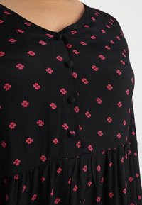 Blouse noire à fleurs avec un décolleté en V, un petit motif de fleurs roses et un design à volants. Se ferme avec trois boutons noirs au niveau du cou.