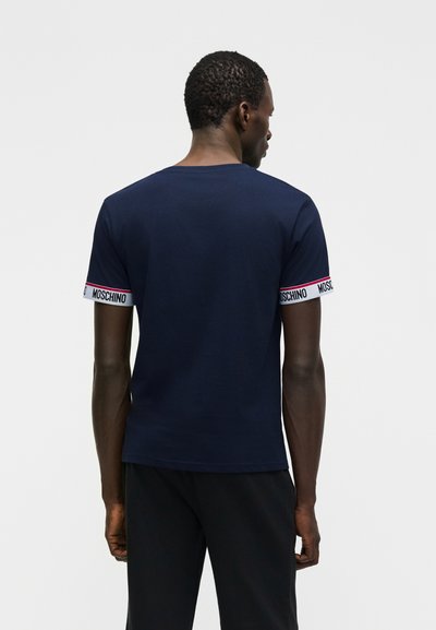 Άνδρας που φοράει μπλε σκούρο κοντομάνικο πουκάμισο με λευκές ρίγες και το κείμενο "Moschino" στα μανίκια, γυρισμένος με την πλάτη προς τα εμάς σε απλό φωτεινό φόντο.