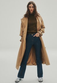 Cappotto trench beige sopra un maglione verde scuro, abbinato a jeans a zampa d'elefante blu scuro e sneakers bianco e nero. Tessuto liscio, vestibilità casual.