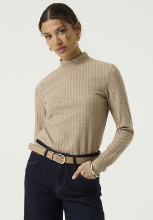 Vrouw met donker haar draagt een beige coltrui, donkere jeans, beige riem, gouden oorringen en een gouden armband, hand in de zak.