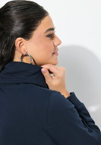 Pullover blu navy con colletto alto, caratterizzato da una trama liscia. Modella con grande orecchini a cerchio intrecciati e che tocca il colletto.