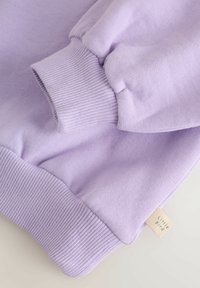 Ljubičasta majica dugih rukava s rebrastim manžetama i rubom. Mekane teksture, s malom naljepnicom "Little Bird" na donjem rubu.