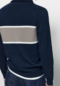 Homme portant un pull bleu marine avec une large bande horizontale grise et un liseré blanc à l'arrière, associé à un jean bleu foncé.
