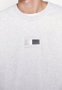 Sudadera gris con una tela texturizada, que presenta un parche de logo rectangular en plateado y negro. Cuello redondo y ribete acanalado.