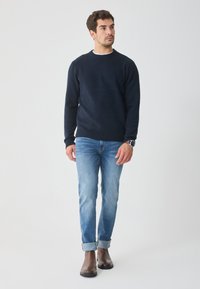 Marineblauer Strickpullover mit rundem Halsausschnitt, gerippten Bündchen, kombiniert mit hellblauen Jeans, die an den Säumen aufgerollt sind, getragen mit braunen Leder-Ankle-Boots.