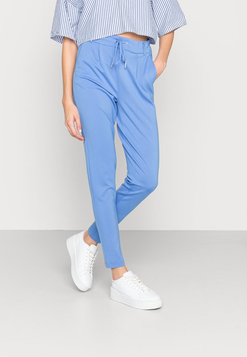 Personne portant un pantalon de jogging bleu clair à cordon, des baskets blanches à plateforme et un haut rayé court, debout contre un mur uni.