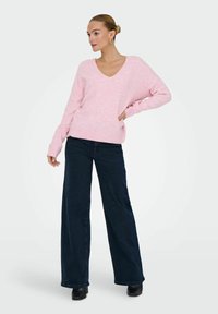 Lichtroze grofgebreide trui met een V-hals, gecombineerd met donkere brede jeans. De trui heeft geribbelde boorden en zoom. Zwarte laarzen met hak.