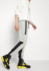 Grå joggerbyxor med svarta sidopaneler och dragkedjefickor, i kombination med svarta och gula sneakers. Avslappnad huvtröja på modellen.