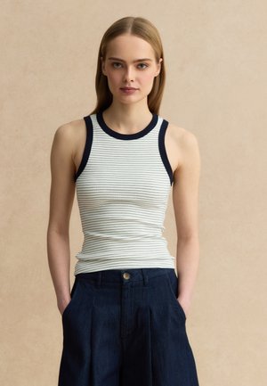 Joven con cabello rubio liso que lleva una camiseta sin mangas blanca acanalada con ribete negro y pantalones de talle alto azul marino.