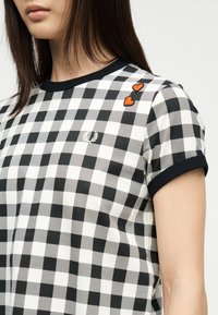 Chemise à manches courtes à carreaux noir et blanc avec bordure noire et deux petites patches de cœurs rouges près de l'épaule sur une femme aux cheveux noirs et lisses.