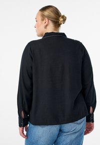 Čierna košeľa s dlhým rukávom a golierom, so zapínaním na gombíky na manžetách a bočnými rozparkami. Mäkká textúra s kontrastnými švy.
