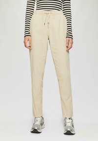 Beige, lichte, losse broek met een elastische tailleband en trekkoord. Gemaakt in combinatie met zilveren hardloopschoenen en een zwart-wit gestreept shirt.