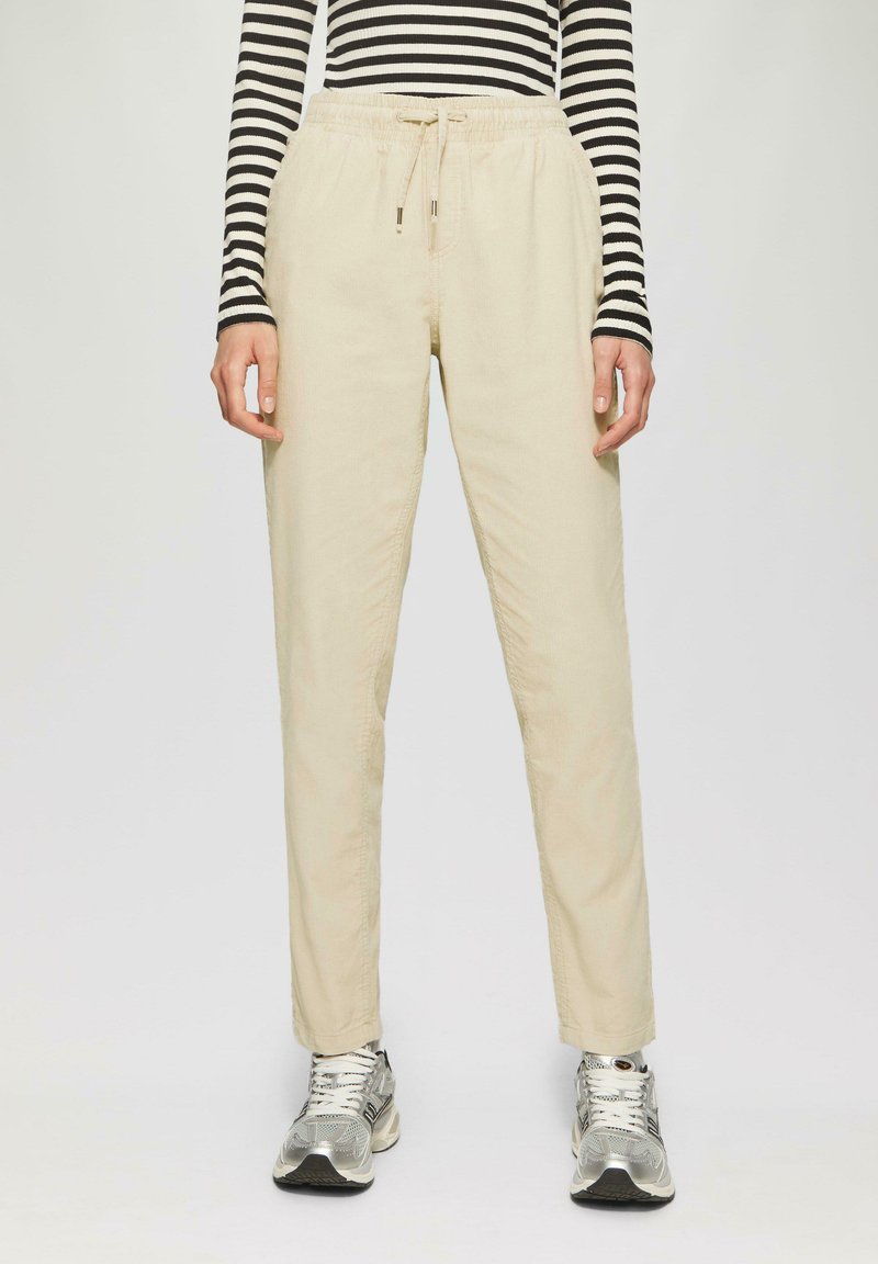 Beige, lichte, losse broek met een elastische tailleband en trekkoord. Gemaakt in combinatie met zilveren hardloopschoenen en een zwart-wit gestreept shirt.