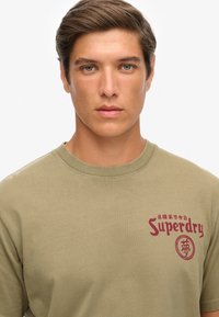 Camiseta de algodón color verde oliva con cuello redondo, que presenta una impresión gráfica en color burdeos de "Superdry" y caracteres japoneses en el lado izquierdo del pecho.